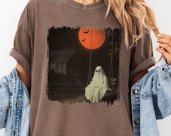 May include: An espresso-colored short-sleeve t-shirt featuring a graphic print of a white ghost sitting on a swing under a large orange moon. The design includes a dark night scene with a house silhouette and two small black bats in the sky.