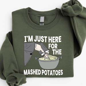 May include: Military green sweatshirt with the text "I'M JUST HERE FOR THE MASHED POTATOES". The graphic shows a person in a suit stirring mashed potatoes in a pot. The brand name "SunsetAndStitchCo" is printed on the top left.