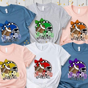May include: A collection of short-sleeved t-shirts in various colors, each featuring a football design with a helmet, bow, and the text "Let's Go Wildcats". The shirts are in shades of blue, pink, gray, and white.