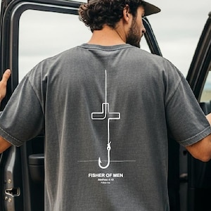 May include: A charcoal gray short-sleeve t-shirt featuring a white line graphic on the back. The design depicts a fishing hook shaped like a cross with the text "FISHER OF MEN", "Matthew 4:19", and "Follow me" printed underneath the hook.