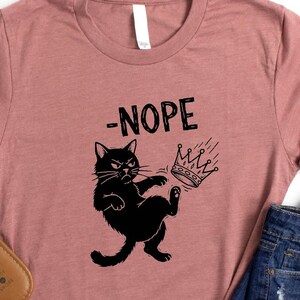 May include: Heather mauve t-shirt with a black graphic of a cat kicking a crown with the word '-NOPE' above it. The shirt is paired with denim shorts and white sandals with a wavy strap design. The SunsetAndStitchCo logo is in the top left corner.