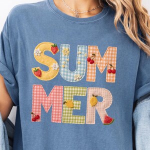 May include: A light blue t-shirt with the word "SUMMER" appliqued on the front. The letters are made of patchwork fabric in various colors and patterns, decorated with fruit and flowers. The text "Blue Jean" is in the top left corner.