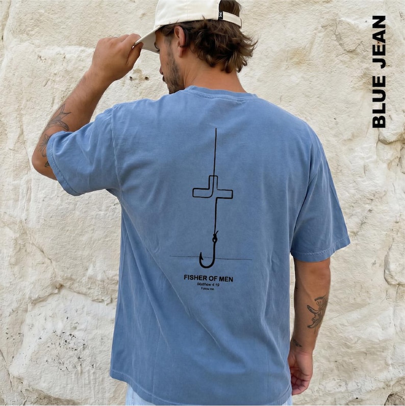May include: A dusty blue short-sleeve t-shirt featuring a black line art graphic on the back. The design depicts a fishing hook shaped like a cross with the text "FISHER OF MEN", "Matthew 4:19", and "Follow Me" printed underneath the hook.
