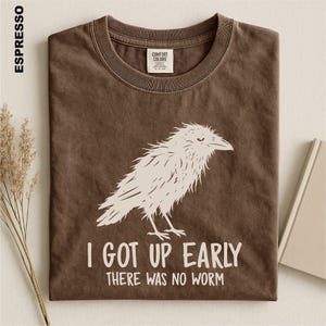 May include: A brown crew neck t-shirt featuring a white graphic of a bird and the text "I GOT UP EARLY THERE WAS NO WORM". The shirt has a Comfort Colors brand label on the inside collar and the color name "ESPRESSO" printed on the left side.