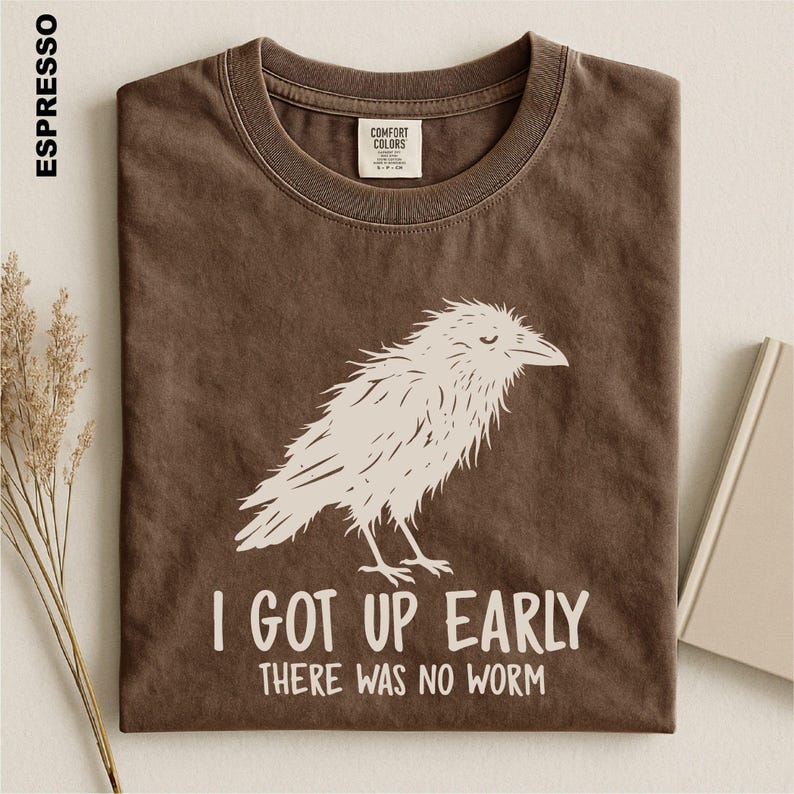 May include: A brown crew neck t-shirt featuring a white graphic of a bird and the text "I GOT UP EARLY THERE WAS NO WORM". The shirt has a Comfort Colors brand label on the inside collar and the color name "ESPRESSO" printed on the left side.