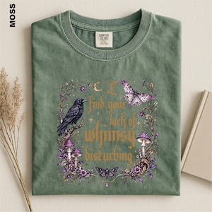 Puede incluir: Una camiseta verde musgo con un estampado gr&aacute;fico de un cuervo negro, setas moradas y mariposas rodeadas por una corona floral. El texto dice, I find your lack of whimsy disturbing. La prenda tiene cuello redondo y una etiqueta en el cuello.