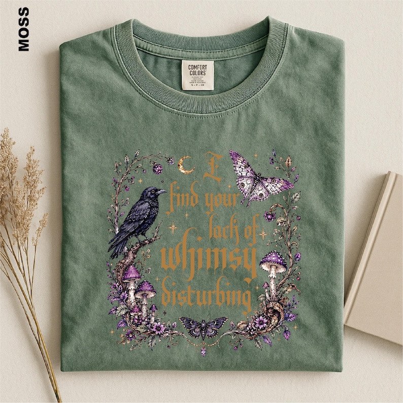 Puede incluir: Una camiseta verde musgo con un estampado gr&aacute;fico de un cuervo negro, setas moradas y mariposas rodeadas por una corona floral. El texto dice, I find your lack of whimsy disturbing. La prenda tiene cuello redondo y una etiqueta en el cuello.