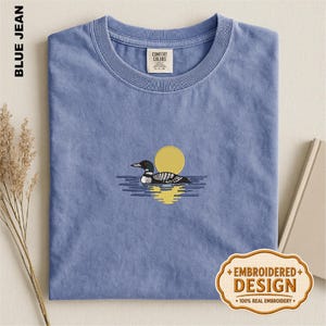 May include: A blue crew neck t-shirt featuring an embroidered design of a loon swimming on water with a yellow sun in the background. The shirt has a Comfort Colors label at the inner neckline and a small tag indicating an embroidered design.