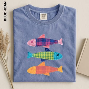 May include: A blue jean colored t-shirt featuring three appliqued fish designs. The top fish is pink and blue plaid, the middle fish is yellow and green gingham, and the bottom fish is orange plaid. Each fish has an X for an eye.