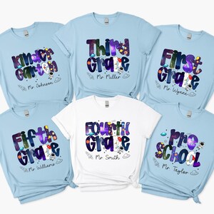 May include: A collection of light blue and white t-shirts with school grade designs. Each shirt features a grade level, such as "Kindergarten" or "Fifth Grade", in a colorful, space-themed font, along with a teacher's name and a rocket ship graphic.