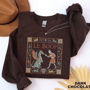 May include: A dark chocolate brown crewneck sweatshirt featuring a graphic print of a medieval knight touching the nose of a green dragon. The text LE BOOP is printed above the figures, all enclosed within a decorative square border.