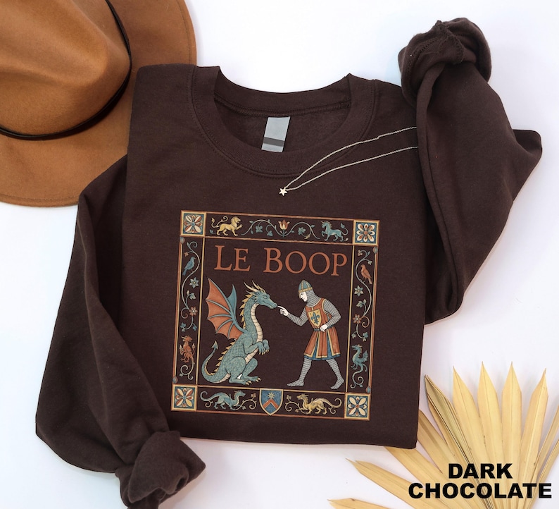 May include: A dark chocolate brown crewneck sweatshirt featuring a graphic print of a medieval knight touching the nose of a green dragon. The text LE BOOP is printed above the figures, all enclosed within a decorative square border.