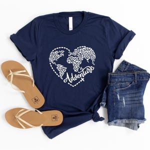 May include: Navy blue t-shirt with a white heart-shaped world map design and the word "Adventure". The shirt is paired with denim shorts and tan sandals. The image is set against a white background.