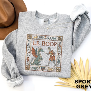 May include: A sport grey crewneck sweatshirt featuring a rectangular graphic with a medieval-style illustration of a knight touching a green dragon's nose. The text LE BOOP is printed above the figures, surrounded by a decorative floral border.