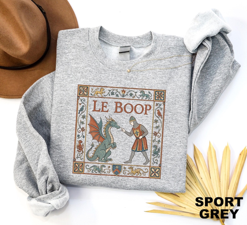May include: A sport grey crewneck sweatshirt featuring a rectangular graphic with a medieval-style illustration of a knight touching a green dragon's nose. The text LE BOOP is printed above the figures, surrounded by a decorative floral border.