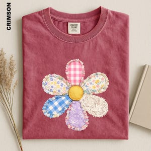 May include: A crimson short-sleeve t-shirt featuring a central flower applique made of mixed fabric patches including pink gingham, blue gingham, floral prints, and white lace. The shirt has a crew neckline and a Comfort Colors brand label.
