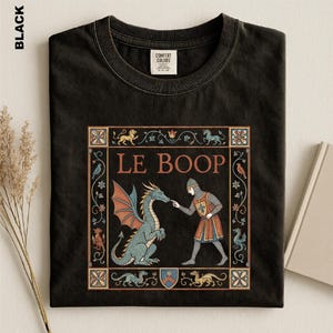 May include: A black t-shirt featuring a medieval-style graphic print with the text LE BOOP. The design depicts a knight in chainmail and a tunic touching the nose of a blue dragon, all framed by a decorative border with floral and animal motifs.