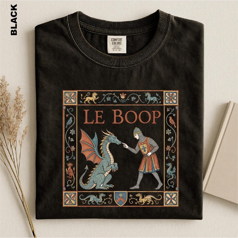 May include: A black t-shirt featuring a medieval-style graphic print with the text LE BOOP. The design depicts a knight in chainmail and a tunic touching the nose of a blue dragon, all framed by a decorative border with floral and animal motifs.