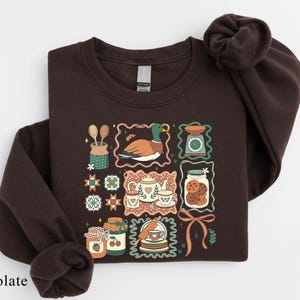 A dark brown sweatshirt with a colorful, illustrated design featuring various kitchen-related items such as spoons, a bird, a cookie jar, and other food-related objects.