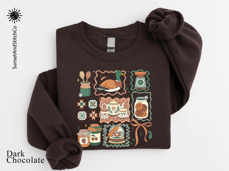 A dark brown sweatshirt with a colorful, illustrated design featuring various kitchen-related items such as spoons, a bird, a cookie jar, and other food-related objects.