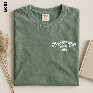 May include: A moss green short-sleeve t-shirt with a crew neckline. The chest features a white graphic print of a fishing hook and line with the text Reel Cool DAD. The shirt has a visible brand label on the inside of the collar.