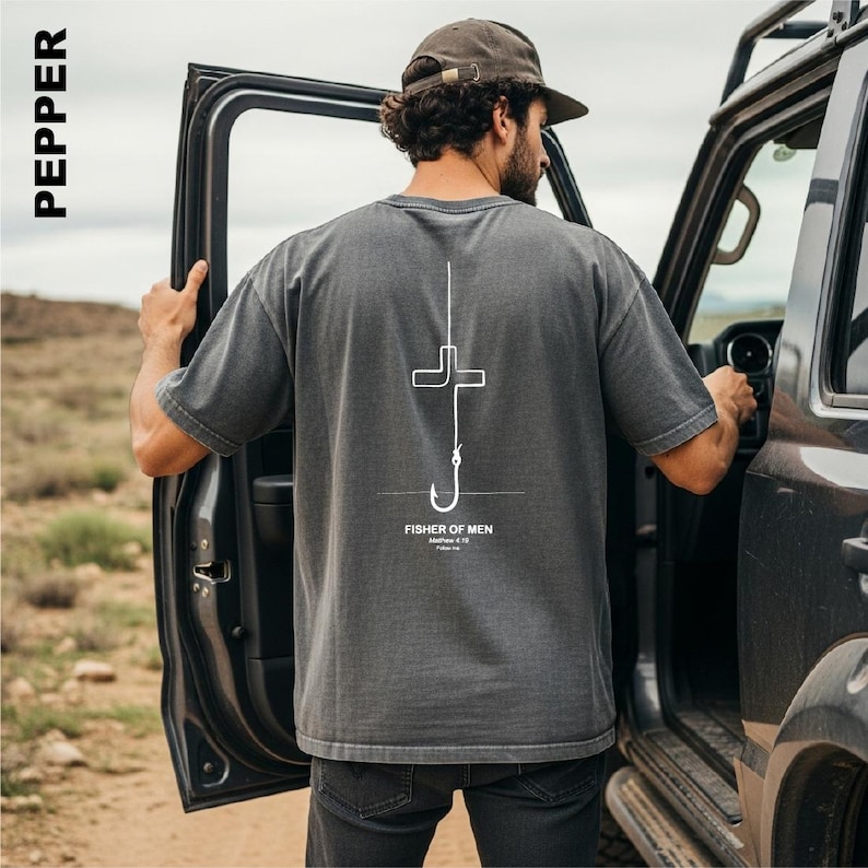 May include: A charcoal gray short-sleeve t-shirt featuring a white line graphic on the back. The design depicts a fishing hook shaped like a cross with the text "FISHER OF MEN", "Matthew 4:19", and "Follow me" printed underneath the hook.