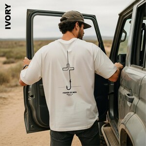 May include: An ivory short-sleeve t-shirt featuring a black line art graphic on the back. The design depicts a cross integrated with a fishing hook. Below the graphic, the text reads "FISHER OF MEN", "Matthew 4:19", and "Follow me".