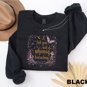 Puede incluir: Una sudadera negra de manga larga con un estampado gr&aacute;fico que incluye el texto "I find your lack of whimsy disturbing". El dise&ntilde;o presenta un cuervo negro, una mariposa morada, setas y estrellas dentro de un marco decorativo en tonos dorados y morados.