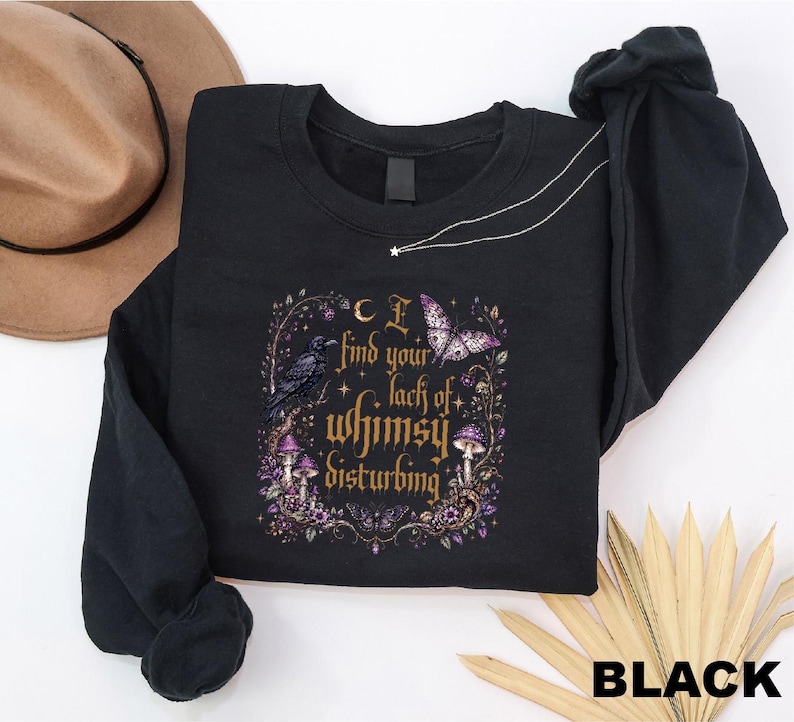 Puede incluir: Una sudadera negra de manga larga con un estampado gr&aacute;fico que incluye el texto "I find your lack of whimsy disturbing". El dise&ntilde;o presenta un cuervo negro, una mariposa morada, setas y estrellas dentro de un marco decorativo en tonos dorados y morados.