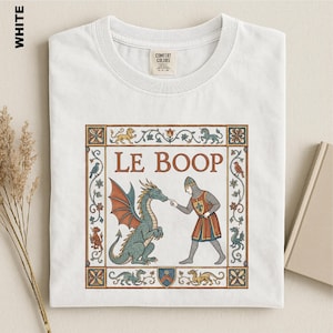 May include: A white t-shirt featuring a medieval-style graphic print. The design shows a knight touching a dragon's nose with the text LE BOOP. The illustration is framed by a decorative border with small mythical creatures and floral motifs.