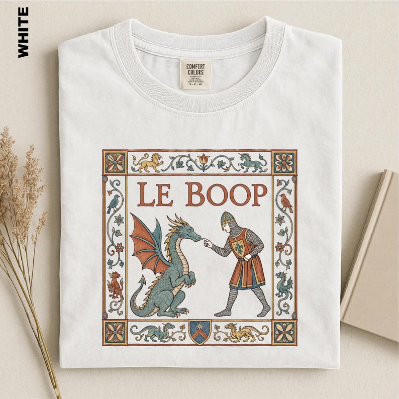 May include: A white t-shirt featuring a medieval-style graphic print. The design shows a knight touching a dragon's nose with the text LE BOOP. The illustration is framed by a decorative border with small mythical creatures and floral motifs.