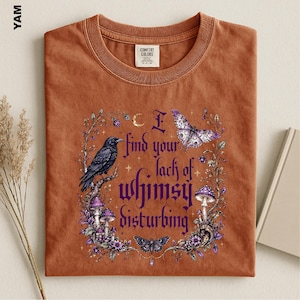 Puede incluir: Una camiseta color &oacute;xido con un estampado gr&aacute;fico de un cuervo negro, setas moradas, mariposas y elementos florales. El texto dice, I find your lack of whimsy disturbing. La prenda tiene cuello redondo y una etiqueta de la marca Comfort Colors.