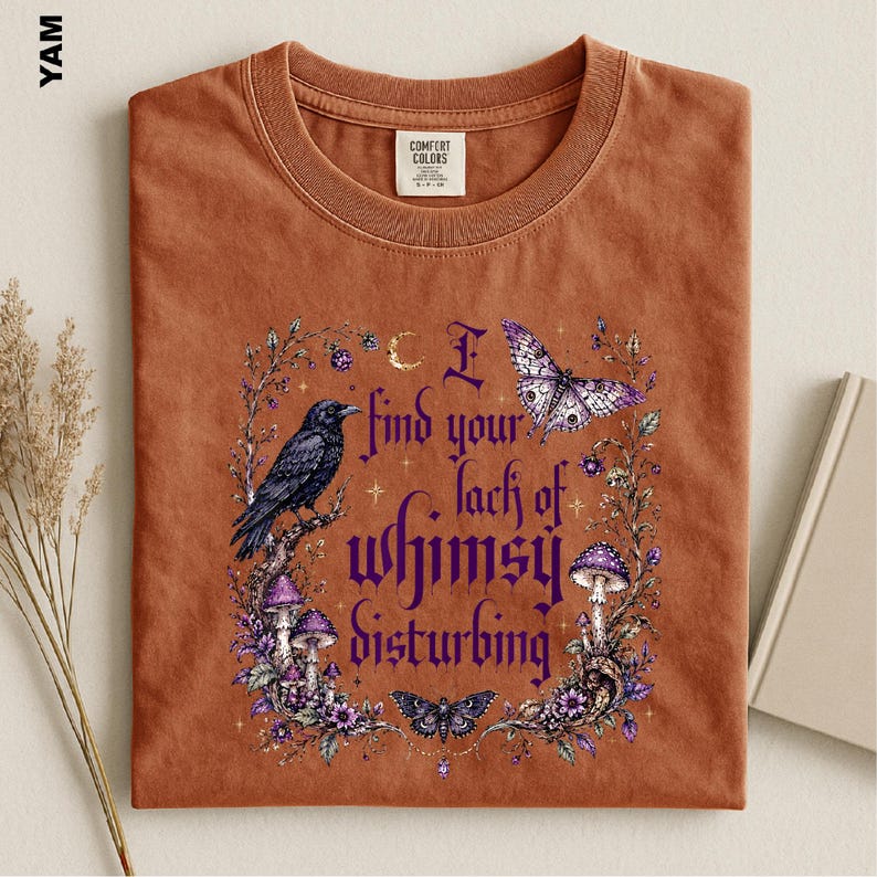 Puede incluir: Una camiseta color &oacute;xido con un estampado gr&aacute;fico de un cuervo negro, setas moradas, mariposas y elementos florales. El texto dice, I find your lack of whimsy disturbing. La prenda tiene cuello redondo y una etiqueta de la marca Comfort Colors.