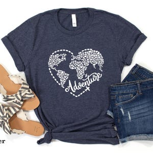 May include: Heather navy t-shirt with a white heart-shaped world map design and the word "Adventure". The shirt is paired with denim shorts and sandals. The text "SunsetAndStitchCo" is in the upper left corner.