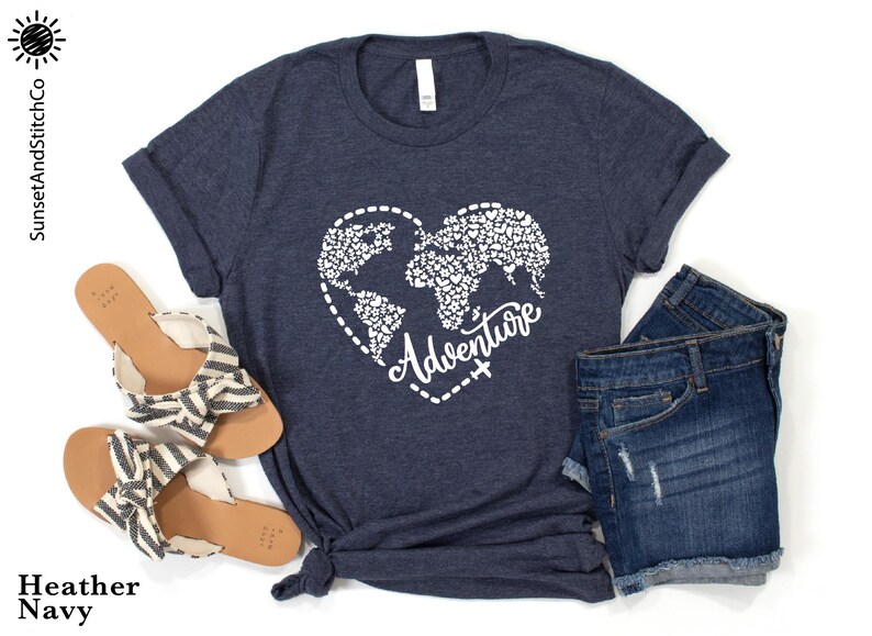 May include: Heather navy t-shirt with a white heart-shaped world map design and the word "Adventure". The shirt is paired with denim shorts and sandals. The text "SunsetAndStitchCo" is in the upper left corner.