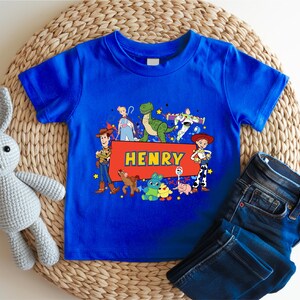 A blue t-shirt with a cartoon character design and the name "Henry" printed on it, along with a pair of jeans and a stuffed rabbit toy.