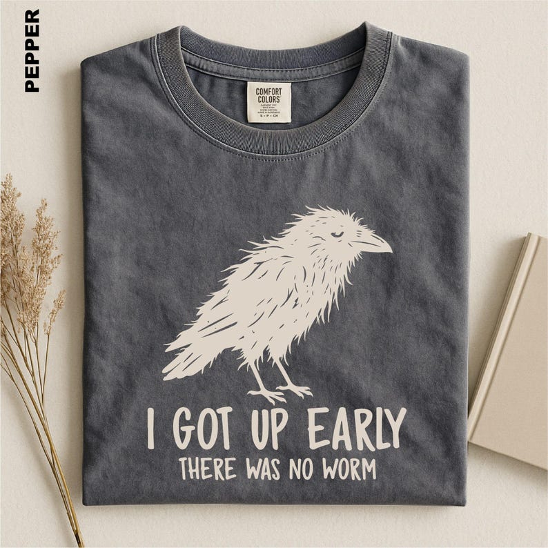 May include: A charcoal gray t-shirt featuring a cream-colored graphic of a bird and the text "I GOT UP EARLY THERE WAS NO WORM". The shirt has a crew neckline and a Comfort Colors brand label on the inside collar. The fabric has a washed appearance.