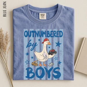 A blue t-shirt with a cartoon image of a duck holding a baseball and a truck, along with the text "OUTNUMBERED BY BOYS" printed on it.