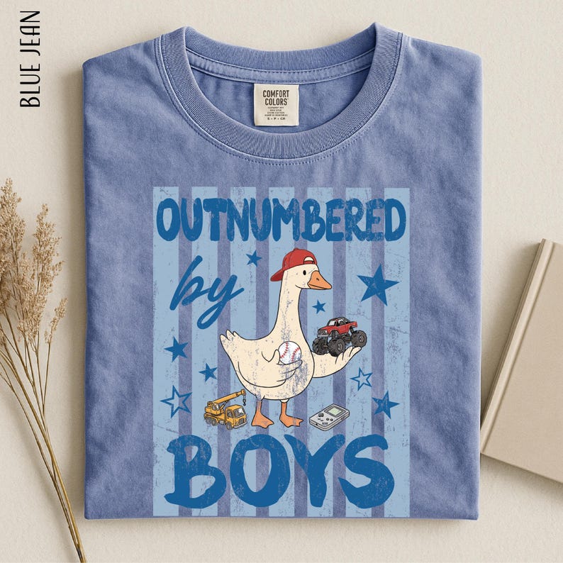 A blue t-shirt with a cartoon image of a duck holding a baseball and a truck, along with the text "OUTNUMBERED BY BOYS" printed on it.
