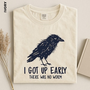 May include: An ivory t-shirt featuring a dark navy blue graphic of a shaggy crow. Below the bird, the text reads: I GOT UP EARLY THERE WAS NO WORM. The shirt has a Comfort Colors brand label at the inner neckline and is displayed flat.