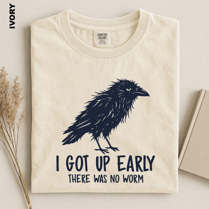 May include: An ivory t-shirt featuring a dark navy blue graphic of a shaggy crow. Below the bird, the text reads: I GOT UP EARLY THERE WAS NO WORM. The shirt has a Comfort Colors brand label at the inner neckline and is displayed flat.