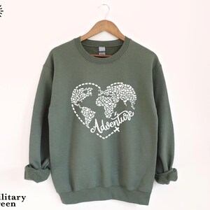 May include: Military green sweatshirt with a white heart-shaped world map design and the word "Adventure". The sweatshirt has long sleeves and a crew neck. The brand name "SunsetAndStitchCo" is printed on the left side.