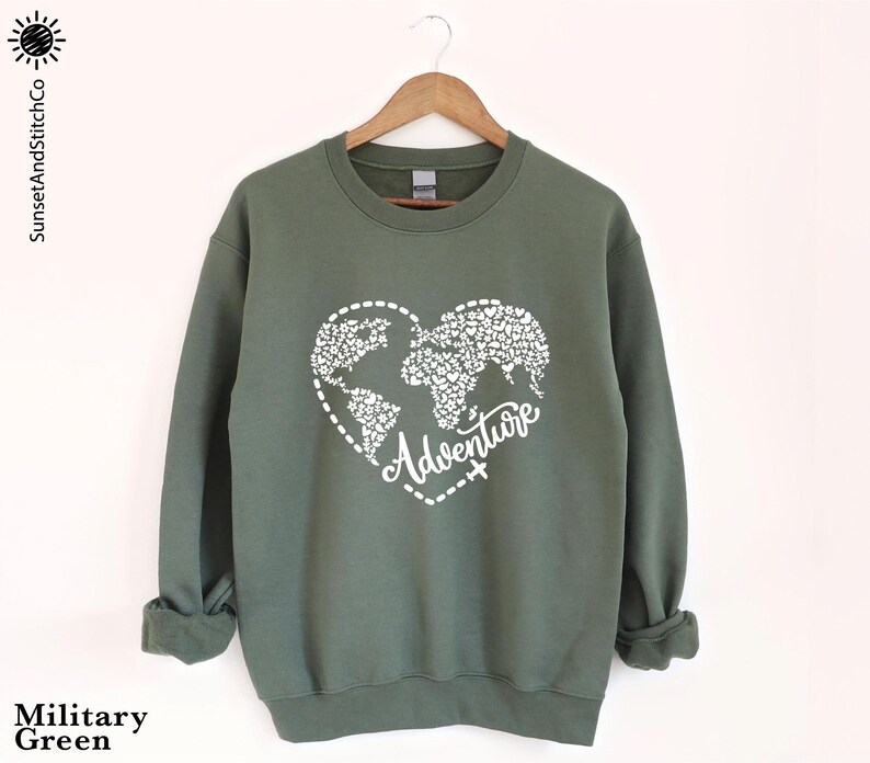 May include: Military green sweatshirt with a white heart-shaped world map design and the word "Adventure". The sweatshirt has long sleeves and a crew neck. The brand name "SunsetAndStitchCo" is printed on the left side.