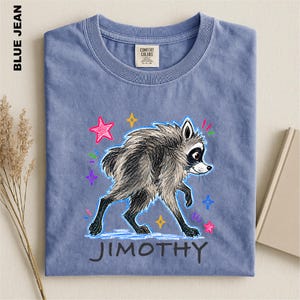 May include: A blue jean colored t-shirt featuring a graphic illustration of a grey raccoon with black markings. The design includes colorful hand-drawn style stars and the name JIMOTHY printed in black capital letters below the raccoon.