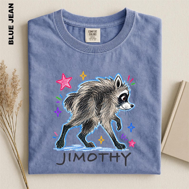 May include: A blue jean colored t-shirt featuring a graphic illustration of a grey raccoon with black markings. The design includes colorful hand-drawn style stars and the name JIMOTHY printed in black capital letters below the raccoon.