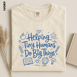 May include: An ivory short-sleeve t-shirt with blue graphic text that reads Helping Tiny Humans Do Big Things. The design includes illustrations of an apple, a star, a crayon, a pencil, an open book, a globe, a lightbulb, and small hearts.