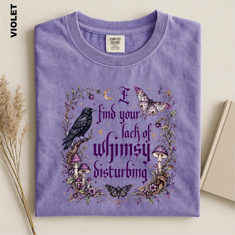 Puede incluir: Una camiseta violeta de cuello redondo con un estampado gr&aacute;fico de un cuervo negro, una polilla, setas y elementos florales. El texto dice, I find your lack of whimsy disturbing. El dise&ntilde;o est&aacute; enmarcado por enredaderas y estrellas en tonos morados y blancos.