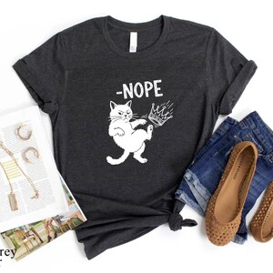 May include: Dark grey heather t-shirt featuring a white cat graphic kicking a crown, with the word "-NOPE". The shirt is styled with denim shorts, brown flats, and a magazine.