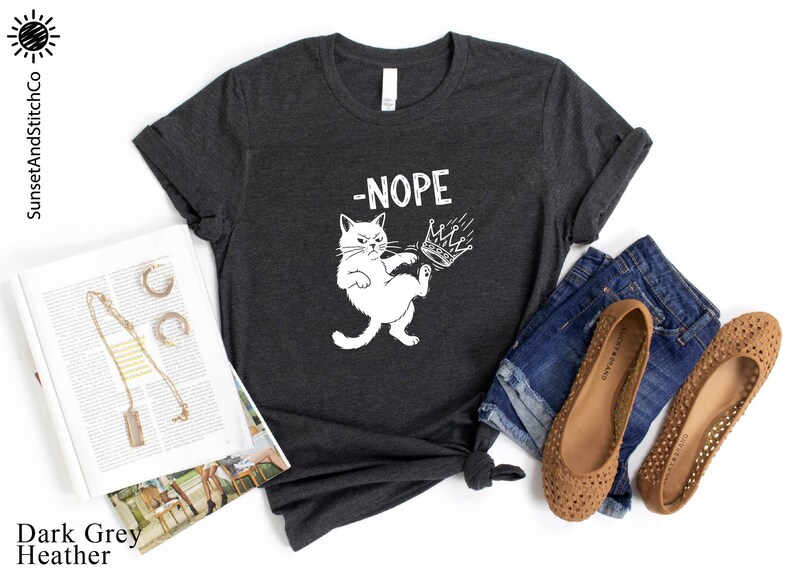 May include: Dark grey heather t-shirt featuring a white cat graphic kicking a crown, with the word "-NOPE". The shirt is styled with denim shorts, brown flats, and a magazine.