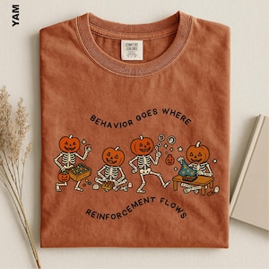 May include: A rust-colored crew neck t-shirt featuring a graphic of four skeletons with pumpkin heads. The text reads "BEHAVIOR GOES WHERE REINFORCEMENT FLOWS" in black lettering. The skeletons are shown engaging in various activities with small toys.